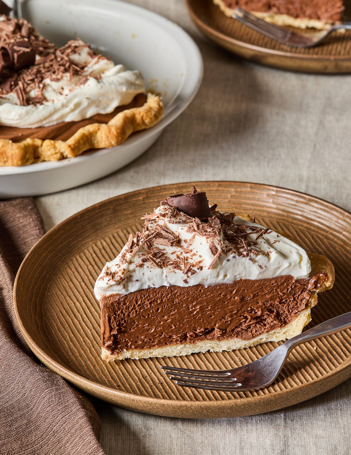 Chocolate pie with whipped cream on a brown plate with a fork, surrounded by other pies.