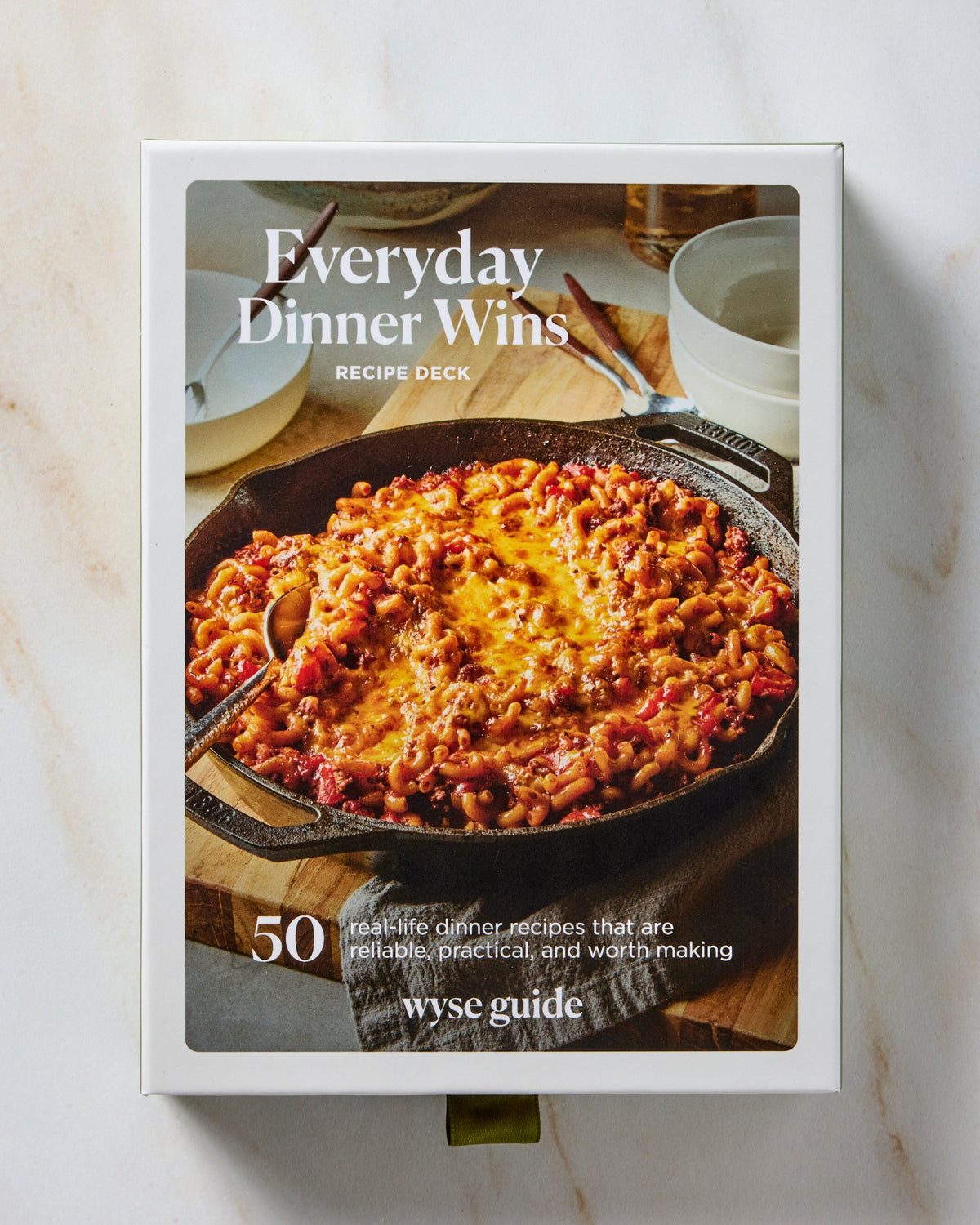 A boxed recipe deck titled "Everyday Dinner Wins" standing upright on a light marble surface. The front cover shows a cast-iron skillet filled with a cheesy baked pasta dish, with text including "Recipe Deck," "50 real-life dinner recipes that are delicious, stress-free, and worth making," and the brand name "wyse guide.