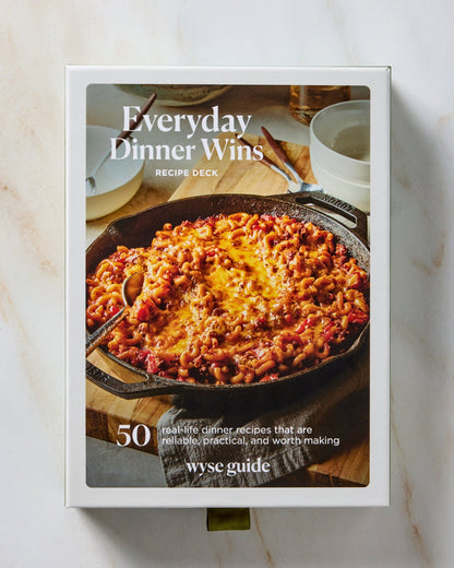 A boxed recipe deck titled "Everyday Dinner Wins" standing upright on a light marble surface. The front cover shows a cast-iron skillet filled with a cheesy baked pasta dish, with text including "Recipe Deck," "50 real-life dinner recipes that are delicious, stress-free, and worth making," and the brand name "wyse guide.