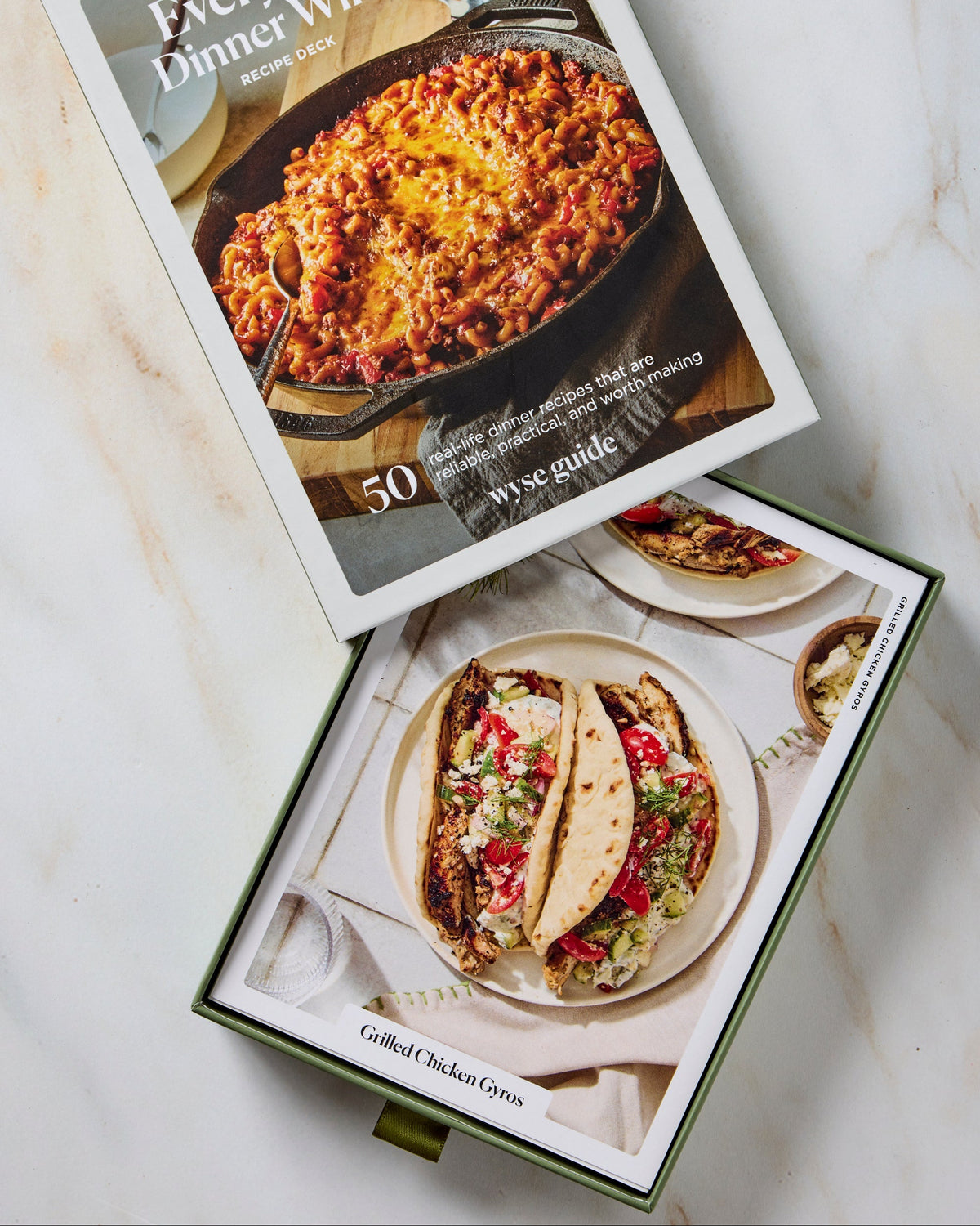 Two boxed cookbooks titled 'Everyday Dinner Wins' and 'Grilled Chicken Tacos' are arranged on a light marble surface. The boxes feature food photography and are photographed from above in a clean, styled flat-lay arrangement.