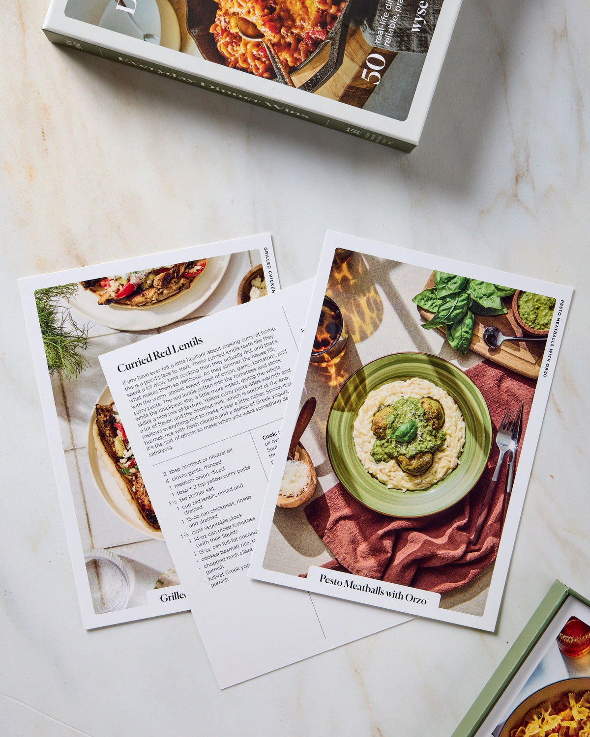 A cookbook titled 'Everyday Dinner Wonders' is shown open on a light marble surface, with one page spread open displaying a recipe for goulash and another showing a plated dish. A few printed recipe cards are scattered around the pages.