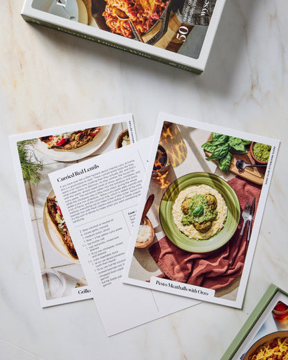 A cookbook titled 'Everyday Dinner Wonders' is shown open on a light marble surface, with one page spread open displaying a recipe for goulash and another showing a plated dish. A few printed recipe cards are scattered around the pages.