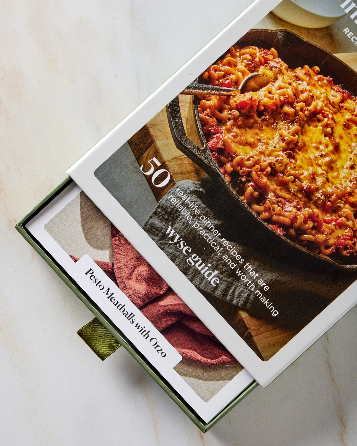 A boxed recipe book or cookbook partially slid open on a light marble surface. The cover shows a photo of a cheesy meatball dish in a cast-iron skillet, with the title '50 Wyse Guide' and a recipe slide for 'Pesto Meatballs with Oreo' visible.