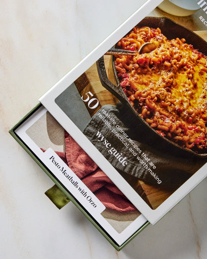 A boxed recipe book or cookbook partially slid open on a light marble surface. The cover shows a photo of a cheesy meatball dish in a cast-iron skillet, with the title '50 Wyse Guide' and a recipe slide for 'Pesto Meatballs with Oreo' visible.