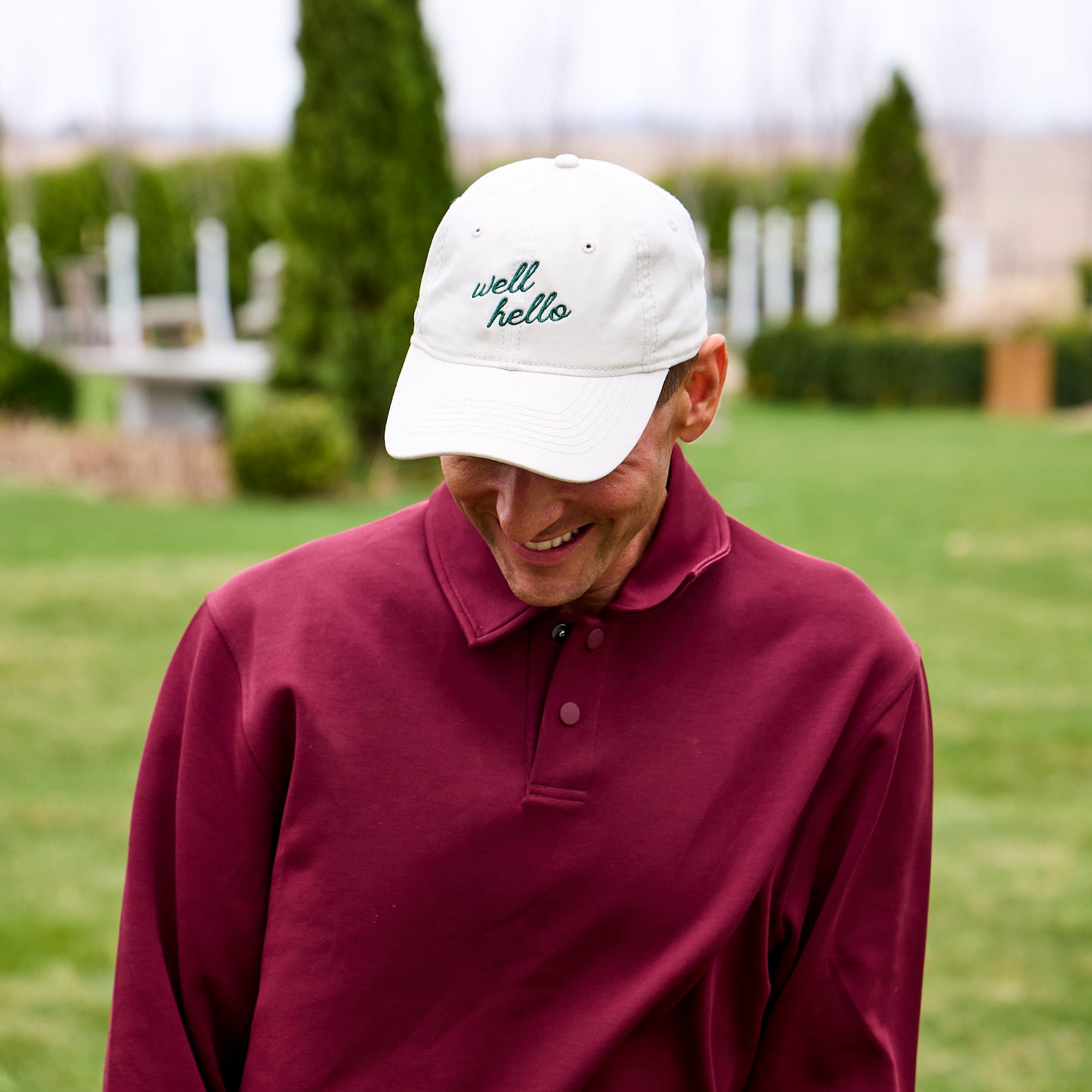 Person wearing a maroon polo shirt and white cap with 'well hello' text outdoors.