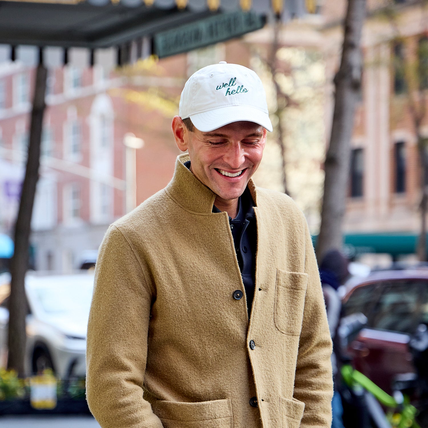 Man wearing a beige coat and white cap with 'well hello' text, standing on a city street.